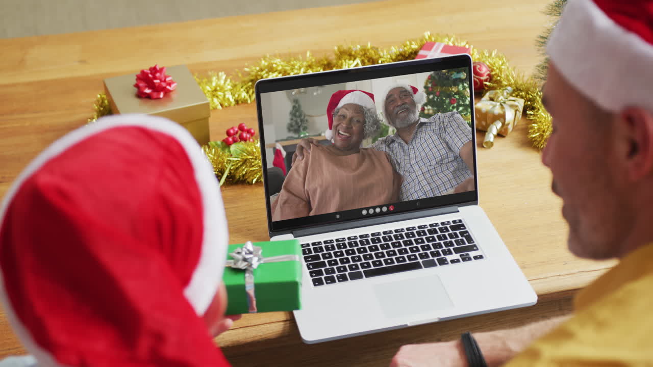 padre caucásico con su hijo usando una computadora portátil para una videollamada de navidad, con la familia en la pantalla