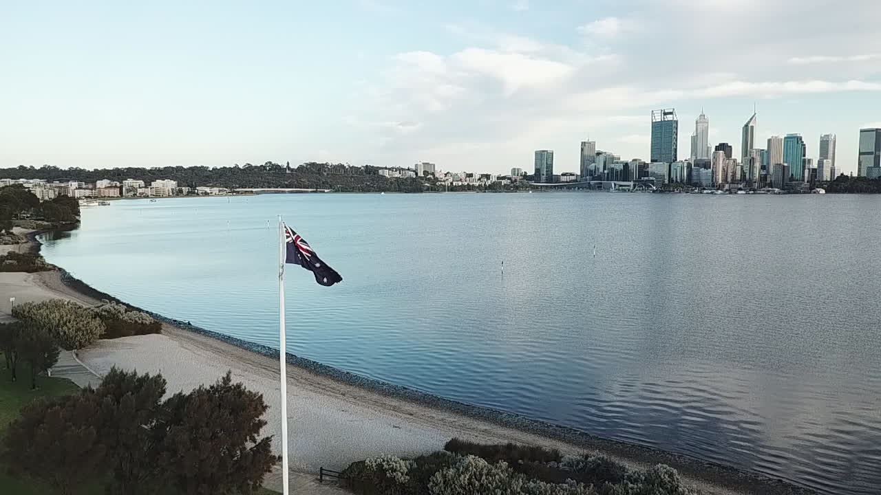 cámara lenta aérea, bandera australiana ondeando en el viento, el río y el horizonte de la ciudad a distancia