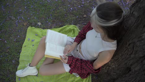 Cute-young-woman-reading-book-sitting-under-tree-in-nature-at-sunset