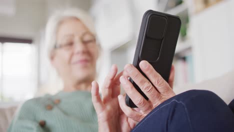mujer caucásica mayor feliz sentada en el sofá de la sala de estar, usando un teléfono inteligente