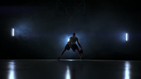 Ein-Mann-Mit-Einem-Basketball-Auf-Einem-Dunklen-Basketballfeld-Vor-Dem-Hintergrund-Eines-Basketballrings-Im-Rauch-Zeigt-Dribbelfähigkeiten,-Beleuchtet-Von-Drei-Laternen-Im-Gegenlicht