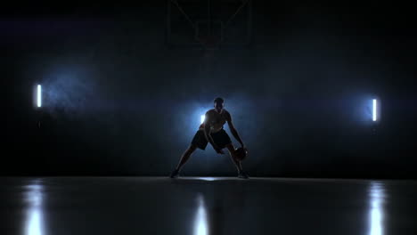 a man with a basketball on a dark basketball court against the backdrop of a basketball ring in the smoke shows dribbling skills illuminated by three lanterns in backlight