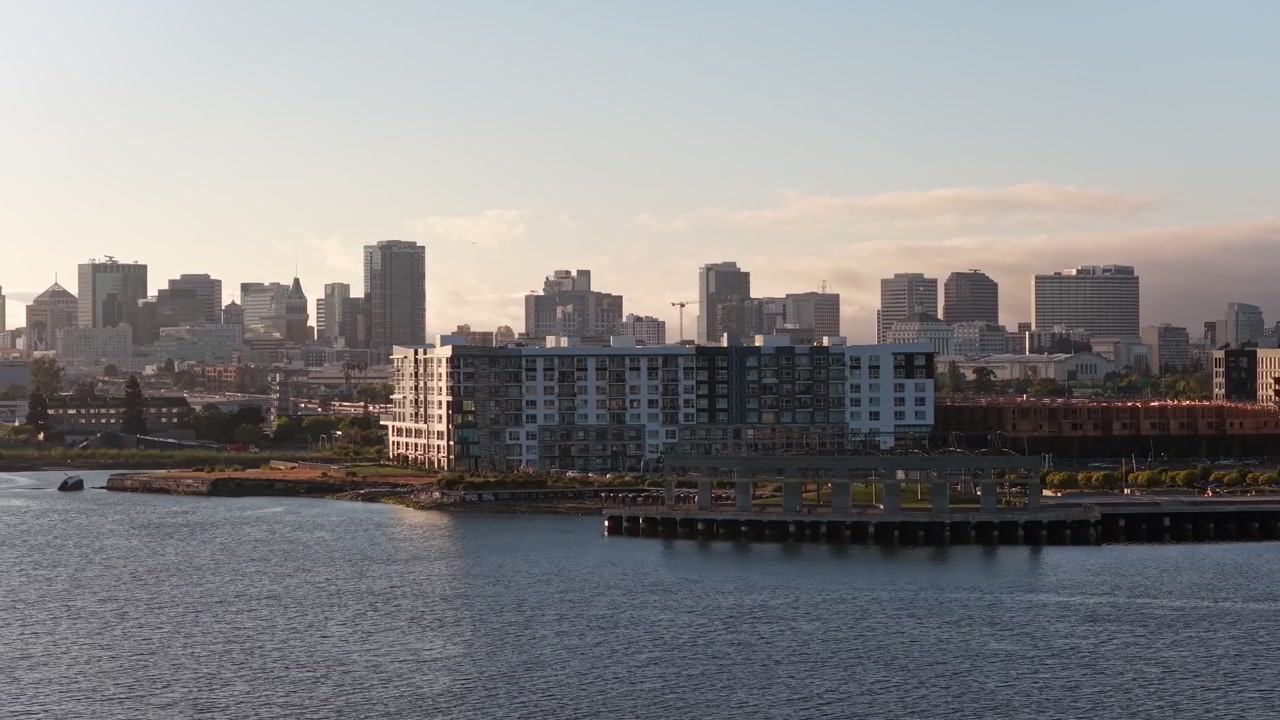 Vídeo aéreo de drone desliza sobre uma orla tranquila, apartamentos modernos e um horizonte urbano vibrante na hora dourada, com luz solar suave e céu aberto criando uma atmosfera urbana pacífica.