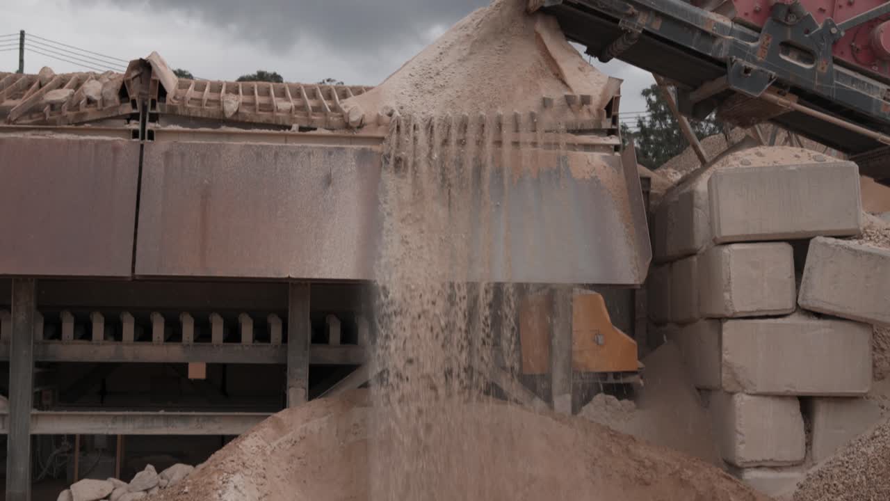 A conveyor system deposits fine sand onto a stockpile at a quarry site. The machinery continuously feeds material, with sand spilling over into collection areas