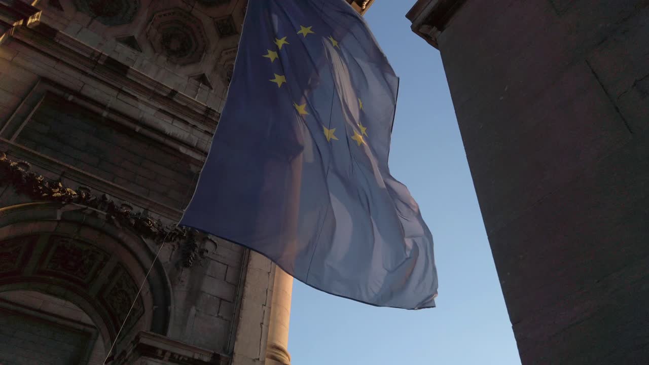 La bandiera dell'Unione Europea sventola con grazia al rallentatore sotto il monumento del Cinquantenario a Bruxelles. Luce dorata e architettura ornata incorniciano questo elegante simbolo di unità e pace.