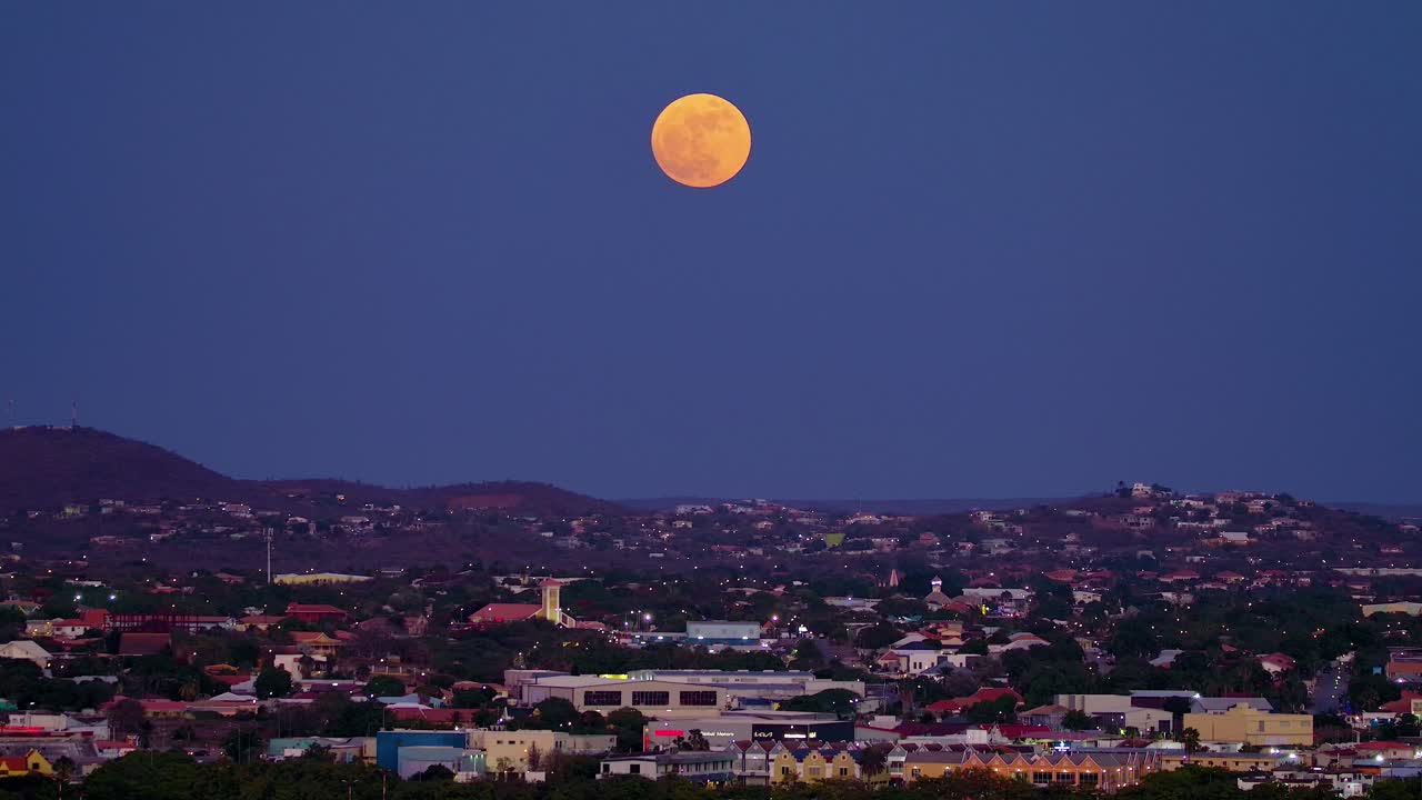 Panorámica hacia arriba a través de la terminal de la zona portuaria industrial de contenedores de Curazao hacia una superluna rojo anaranjado sobre la ciudad de Willemstad