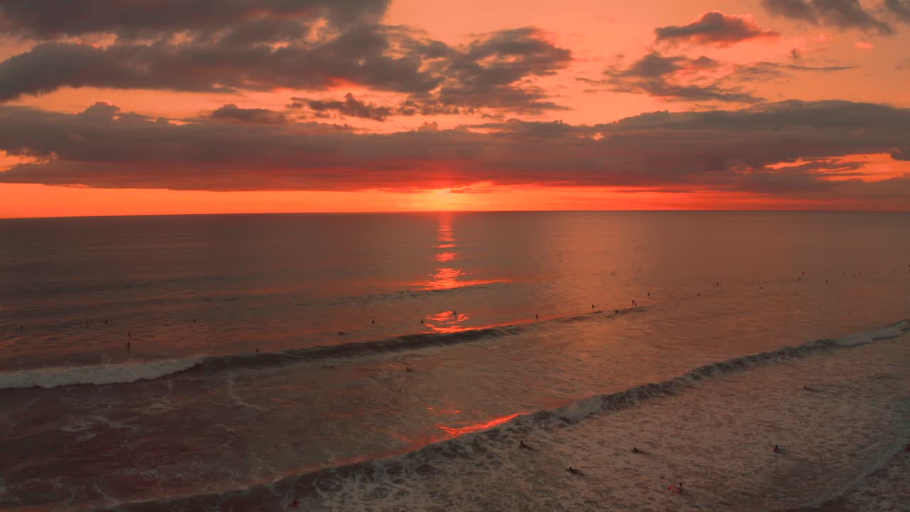Epic aerial shot of a wide sandy beach at the Pacific Coast in Costa Rica at sunset with a beautiful colored sky and clouds above the ocean. Surfers are riding the waves. Shot in 4K.