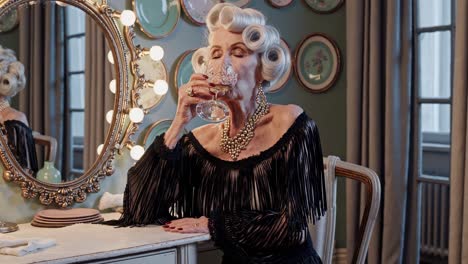 glamorous senior woman with styled hair and elegant black dress enjoys a glass of wine at her vintage vanity table, savoring the moment in a luxurious setting