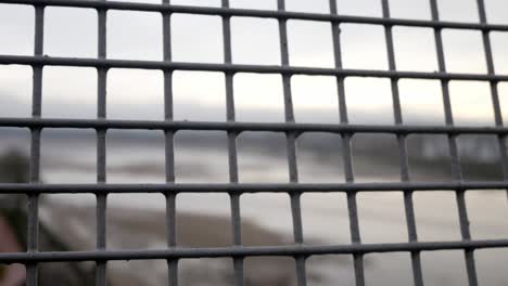 Industrial-grid-security-mesh-fence-barrier-closeup-overlooking-blurred-bokeh-waterfront-high-bridge-view-dolly-right