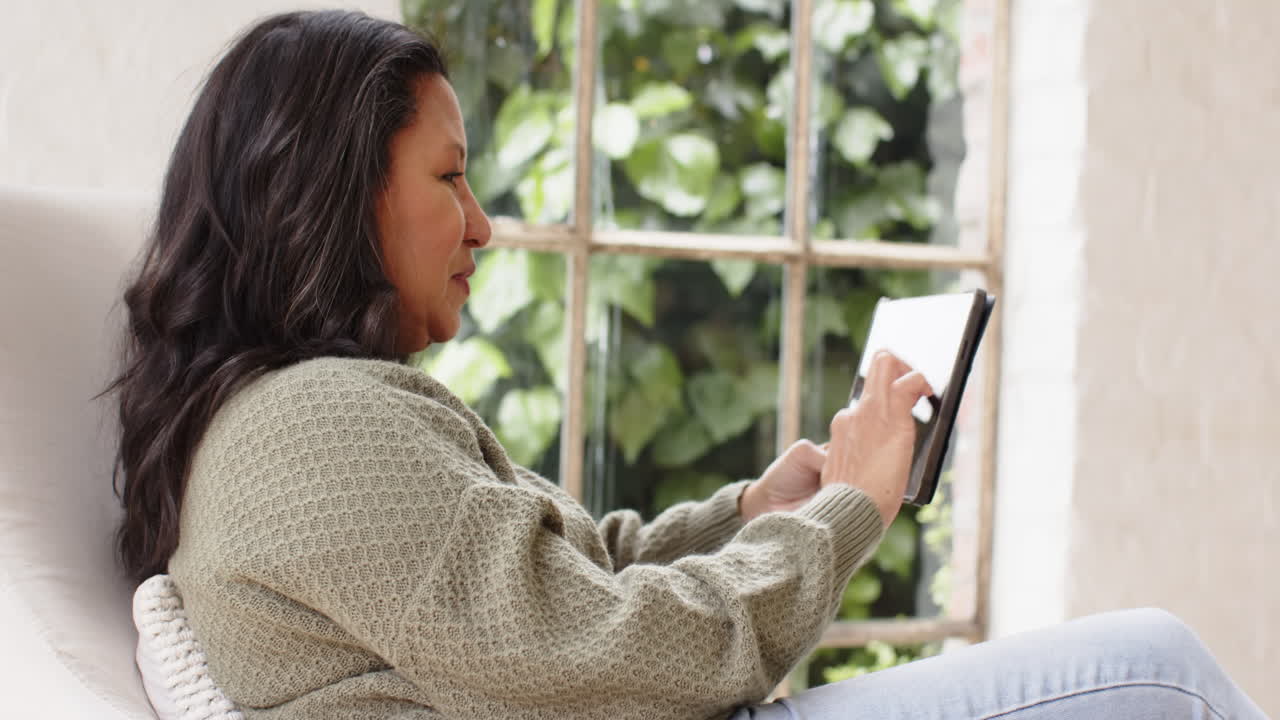 Senior woman relaxing at home using tablet near window, enjoying leisure time