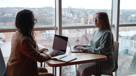business ladies discussing ideas, building strategies, cooperating. women sit in front of each other having laptops on the desk. panoramic window at backdrop.