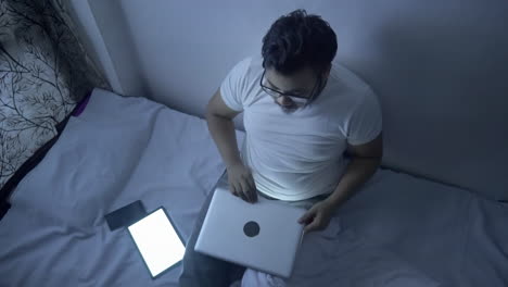 top view of a man sitting in front of a computer screen for long time at night on bed, feeling tired from his work