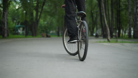 rear view of person in sneakers standing while riding a bicycle on paved road in an irregular manner, captured from a low angle, in the background, another person sits on a bench