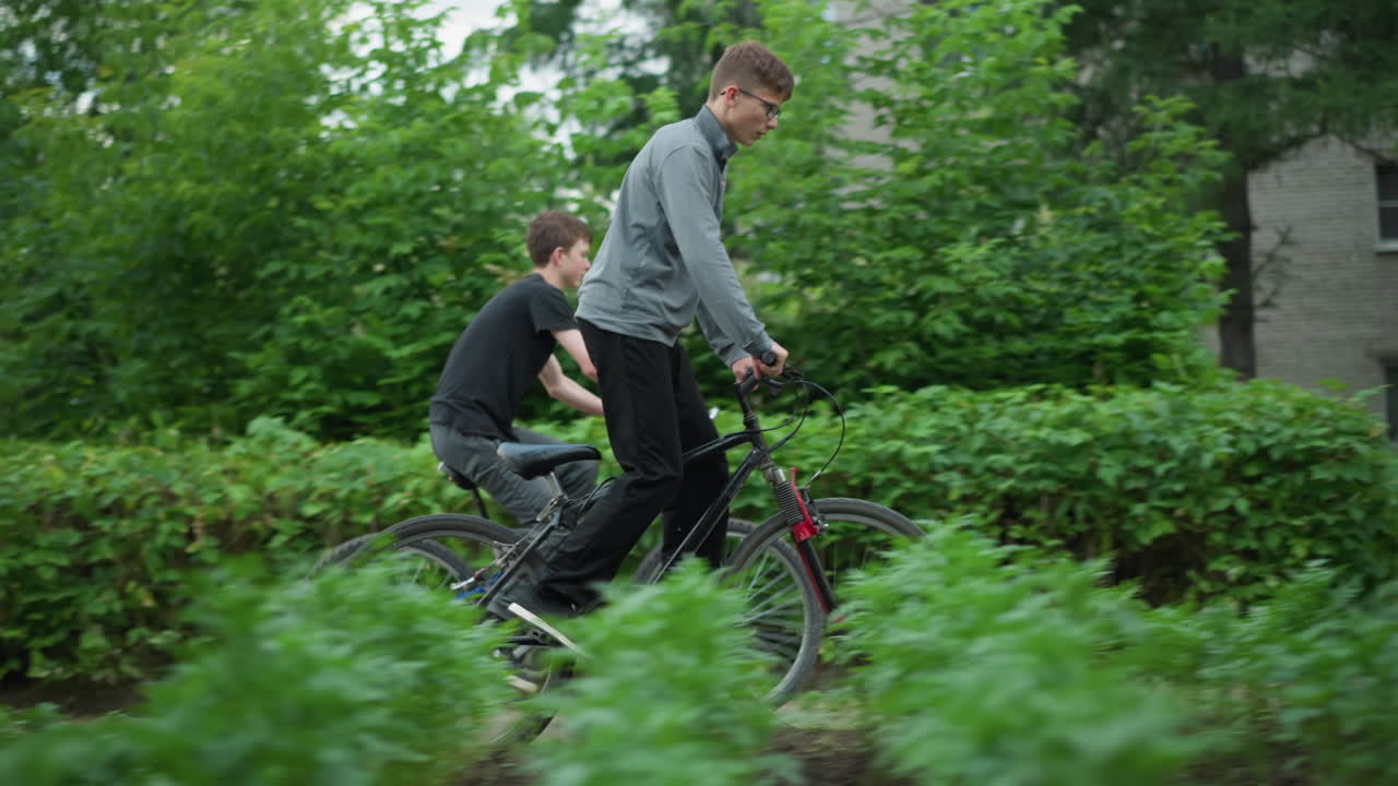 두 친구가 울창한 초록색 지역을 가로질러 자전거를 타고 있는 측면 전망, 한 명이 회색 에 서 있고, 다른 한 명이 검은 에 앉아 있으며, 배경에는 초록색, 흐릿한 건물이 있습니다.
