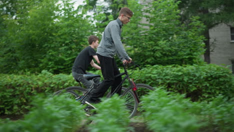 side view of two friends riding bicycles through a lush green area, one in a grey top standing while riding, and the other in a black top seated, the background features greenery, blurred buildings