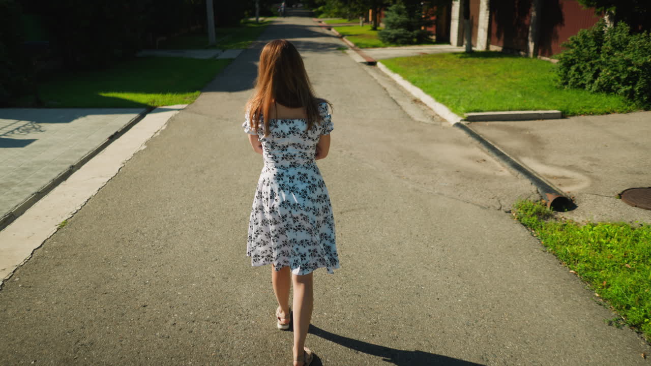 Back view of young woman in floral dress walking thoughtfully with folded arms down quiet residential street in bright daylight, casting long shadow on pavement near green lawn and suburban homes