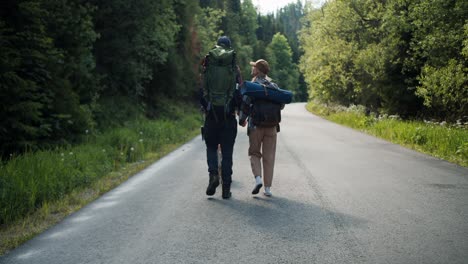 Rear-view-of-a-guy-and-a-girl-tourists-in-special-clothes-for-hiking-with-large-backpacks-walk-along-the-road-along-the-green-forest