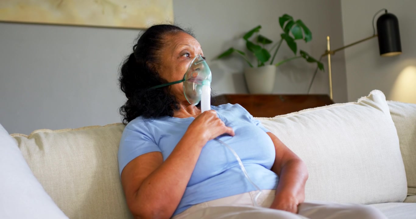 Anziana donna afroamericana che porta una maschera per l'ossigeno al viso, regolando il tubicino, respirando sul divano di casa
