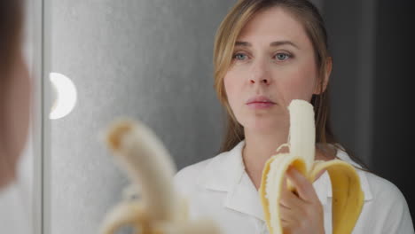 close up rear view of lady eating banana while mirror reflects her expression and action under soft indoor lighting creating calm ambiance as she stands in front of vanity in white shirt enjoying snack