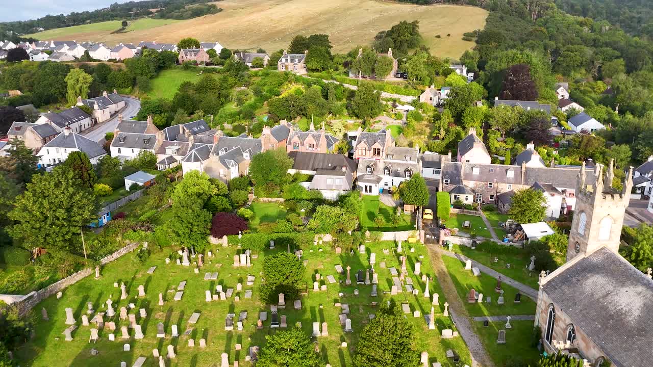 Drone footage glides above a stone church, clock tower, and graveyard surrounded by lush gardens and traditional village buildings in the Scottish Highlands, under bright daylight