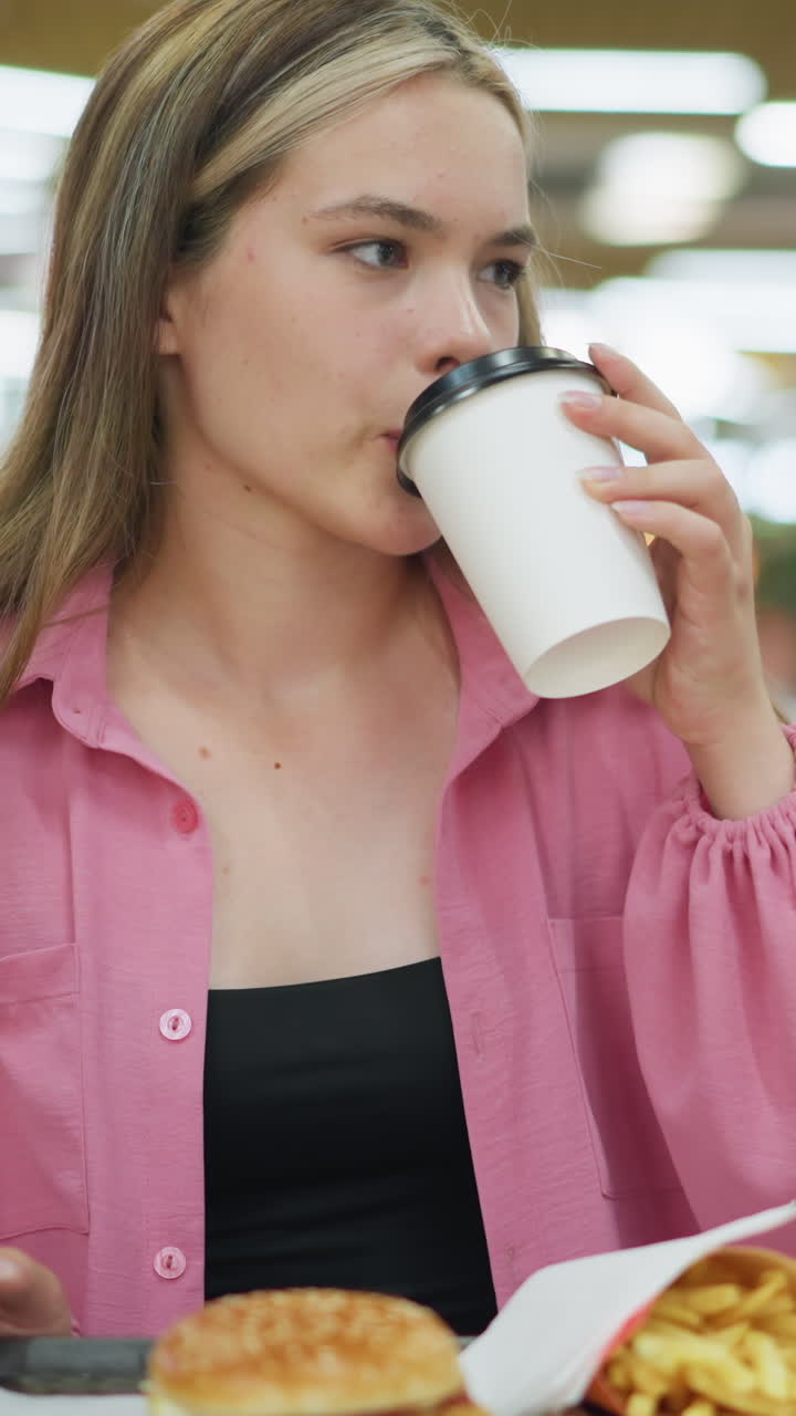 dama de vestido rosa recoge su bebida, toma un sorbo con una expresión satisfecha, y lo coloca de nuevo en la mesa, papas fritas y hamburguesa se colocan en una bandeja, con una vista borrosa de la gente en el fondo