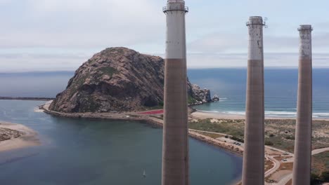 Aerial-parallax-panning-shot-of-the-three-smokestacks-above-the-derelict-Morro-Bay-Power-Plant-with-Morro-Rock-in-the-distance-in-Morro-Bay,-California