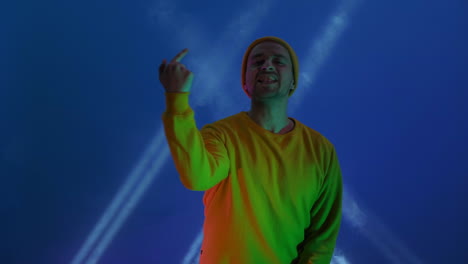 The-man-dances-merrily-and-looks-at-the-camera-in-the-light-of-strobe-lights-and-colored-spotlights.-Neon-colors-and-crazy-man-dancing-to-music-and-singing-in-a-yellow-jacket-and-hat