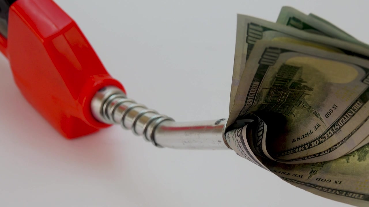 Notas de cem dólares sendo despejadas no tanque de combustível de um veículo, simbolizando o alto custo da gasolina e o peso financeiro de abastecer um carro