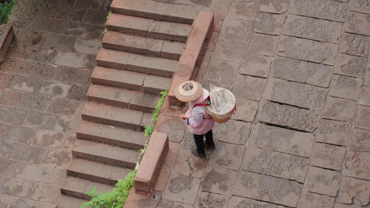 Aerial view woman straw hat walking along cobblestone steps carrying woven basket, vintage brick stairway with greenery edging, textured pavement and moss, overcast light creating quiet market