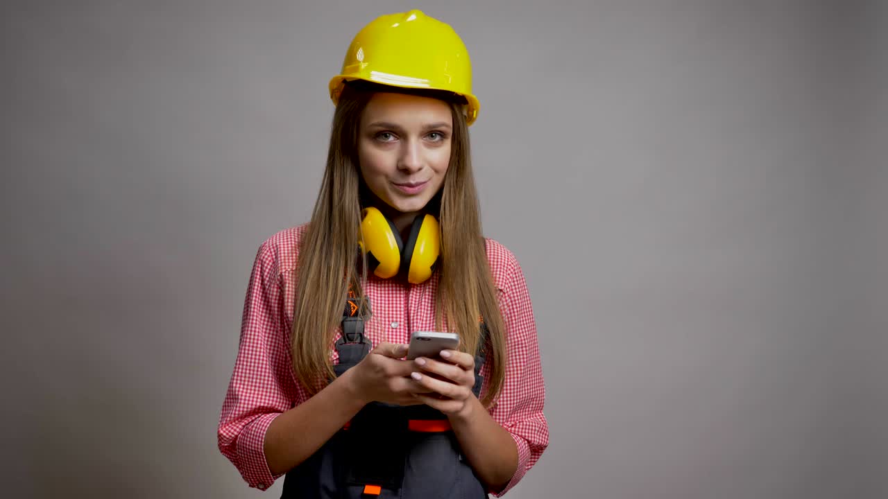 joven constructora bonita está mostrando pantalla croma de su teléfono, sonriendo, concepto de comunicación, fondo gris
