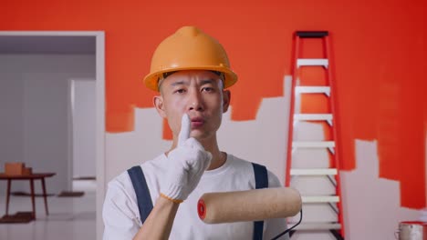 close up of asian man painter wearing safety helmet looking at camera and making shh gesture while standing with painting wall in room
