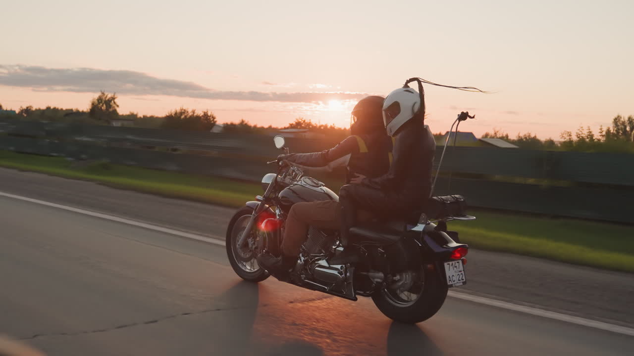 Couple wearing helmets rides motorcycle along open road during sunset, passing greenery and buildings while enjoying road trip adventure, symbolizing unity, freedom and lifestyle