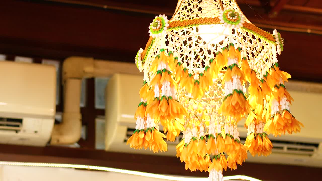 A vibrant Thai flower mobile hangs indoors, illuminated warmly. The intricate design and bright colors create a lively atmosphere