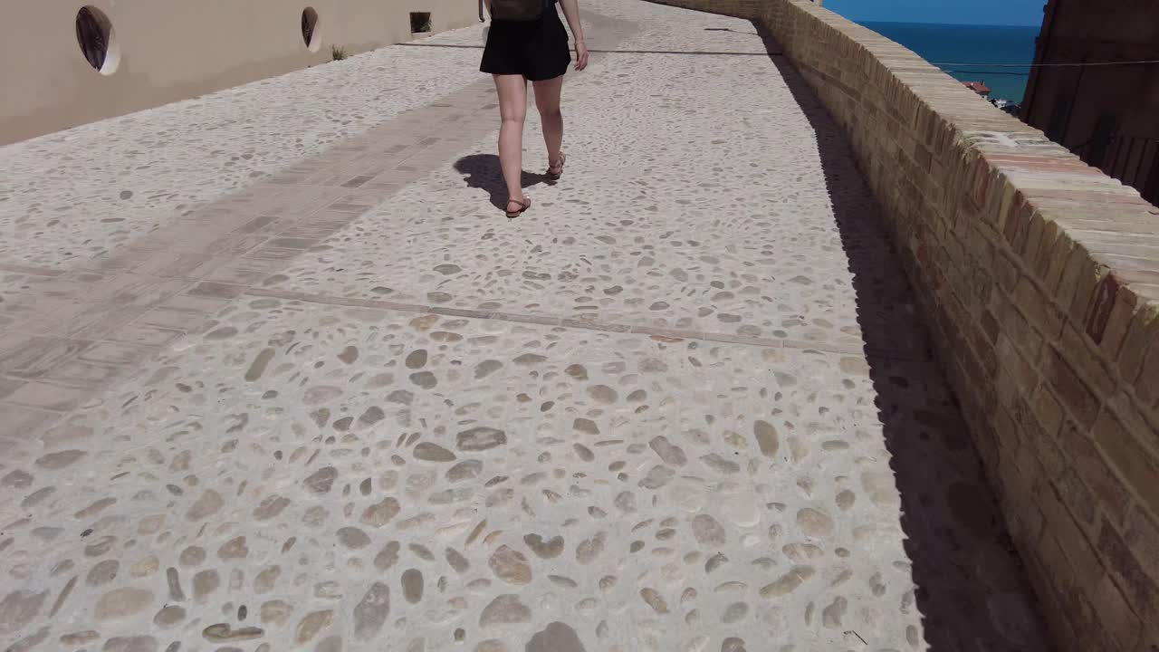 Woman walking on a stone path in the village of Grottammare in Italy. Ancient buildings and on the horizon the blue sky and the sea.