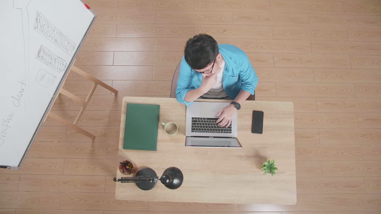 뷰  아웃 : 아시안 남자가 집에서 화이트보드 의 나무 책상에 앉아서 스마트 폰 에 노트북으로 생각하고 타이핑하는 것을 위해 을 들고 있습니다.