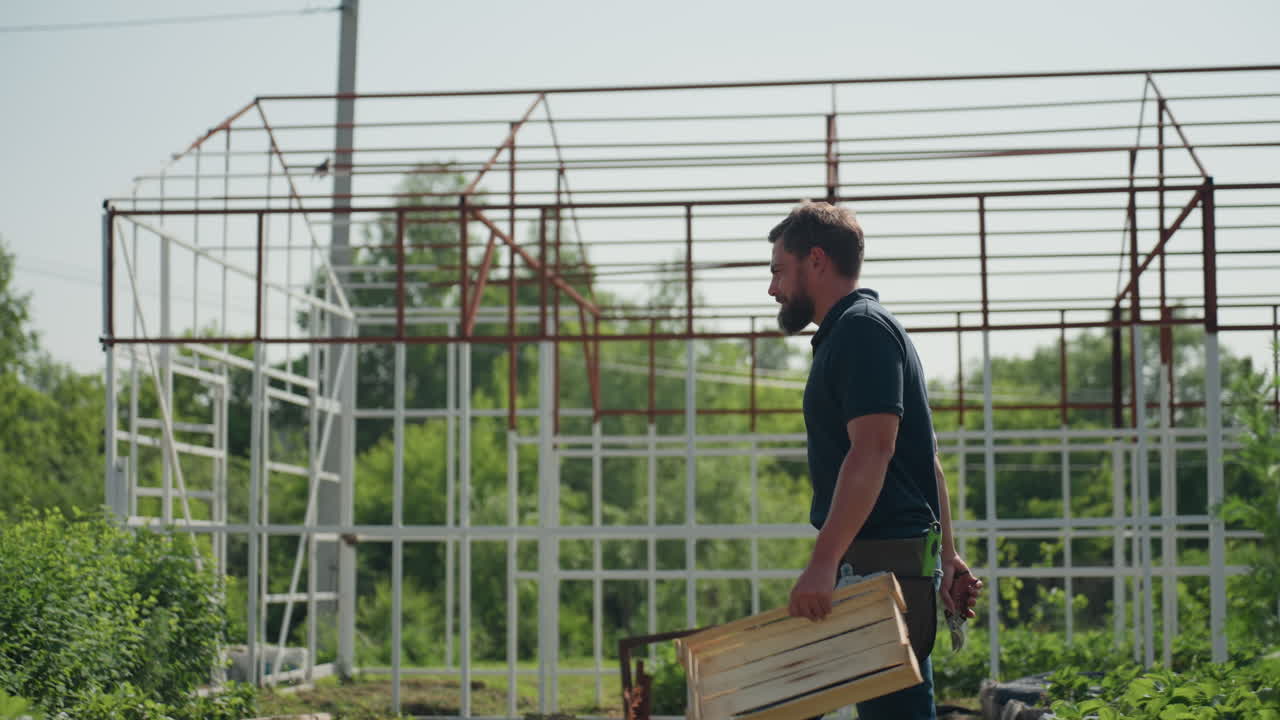 close up green leaves in foreground with side view of horticulturist walking past metal frame structure carrying crate and plier while rubbing nose during plant inspection on sunny rural field