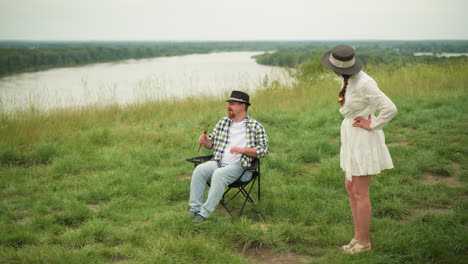 an artist in a checked shirt holds brushes while sitting on a chair in a grassy field beside a lake, demonstrating a sitting pose. a woman in a white dress stands with her hands on her hips, observing