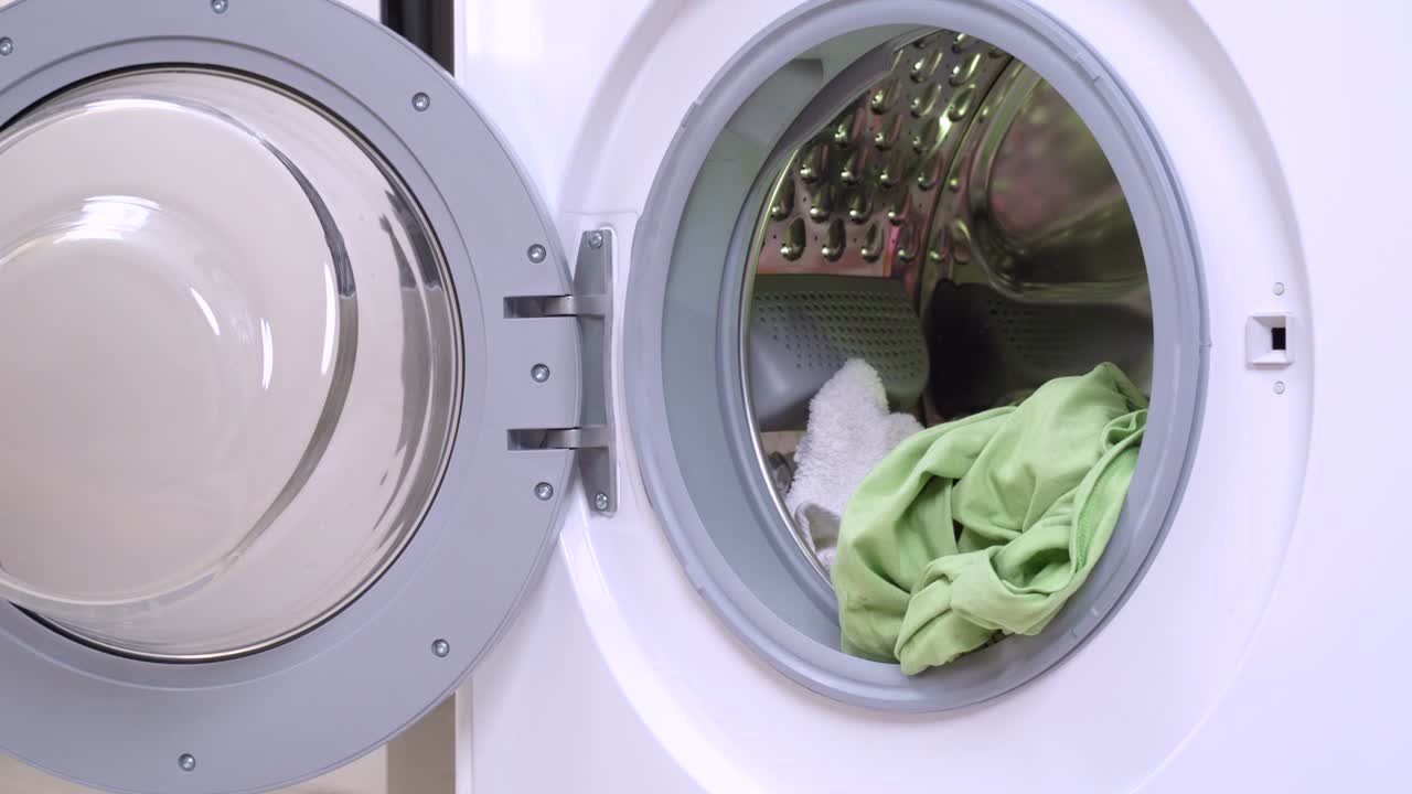 Little black and tan dachshund wearing funny pink and white checkered housemaid costume and golden blond wig uploads laundries into opened washing machine drum. Humor concept of pet housekeeping.