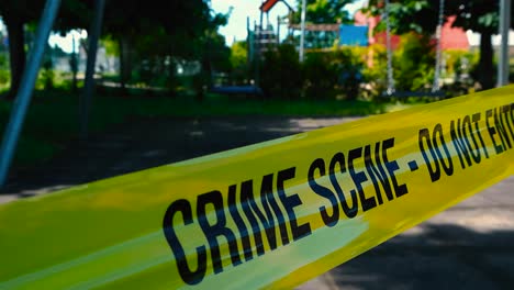 static view of yellow crime scene tape, do not enter in foreground, sunny day. in blurred background closed playground park, empty moving swings. crime against child, kid or children or missing person