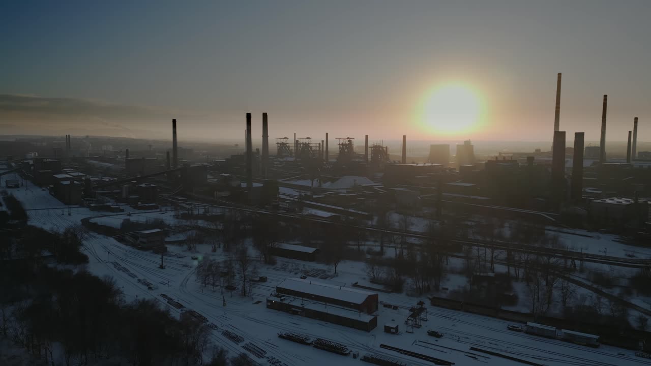 воздушный вид промышленной зоны, покрытой снегом, с высокими, темными дымоходами, силуэтами на живом небе во время захода солнца