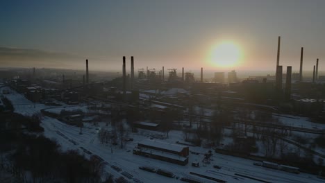 Luftaufnahme-Eines-Schneebedeckten-Industriegebiets-Mit-Hohen,-Dunklen-Schornsteinen,-Die-Sich-Bei-Sonnenuntergang-Vor-Einem-Lebhaften-Himmel-Abheben