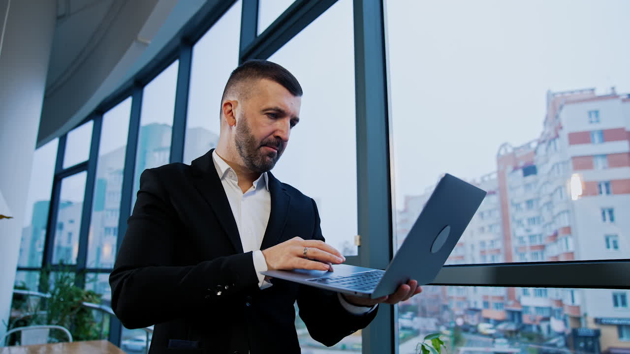 Mid-aged businessman stands at the window holding laptop. Man finishes his work on computer, closes it and looks thoughtfully in the window. Low angle view.