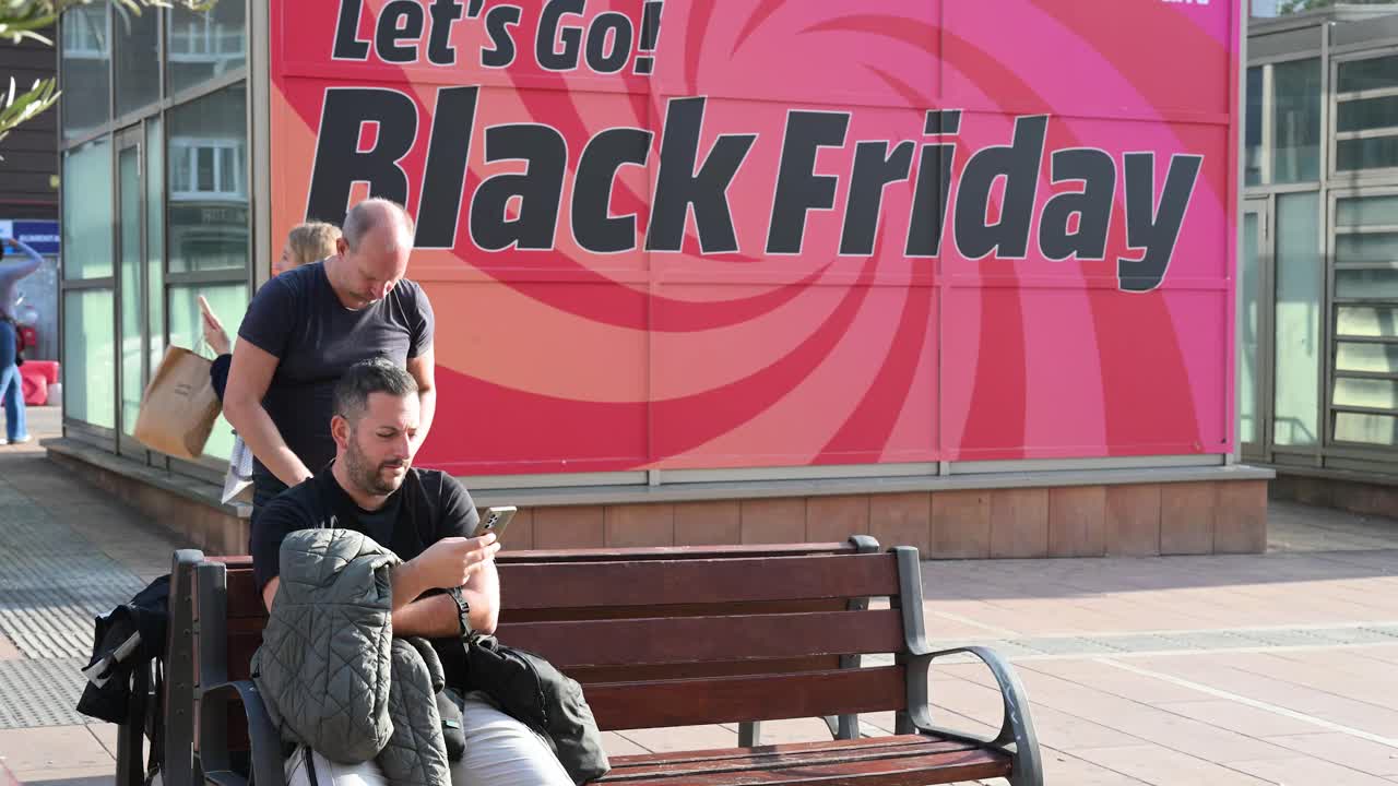 A man sits on a bench in Madrid, Spain, using his smartphone, with a banner advertising Black Friday sales and early holiday deals in the background.