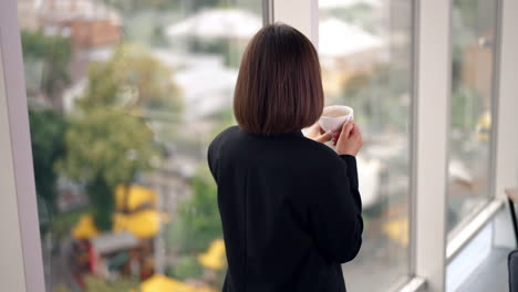 good-looking brunette lady drinking coffee in the office. lady having coffee break at work and looks at window. blurred backdrop.