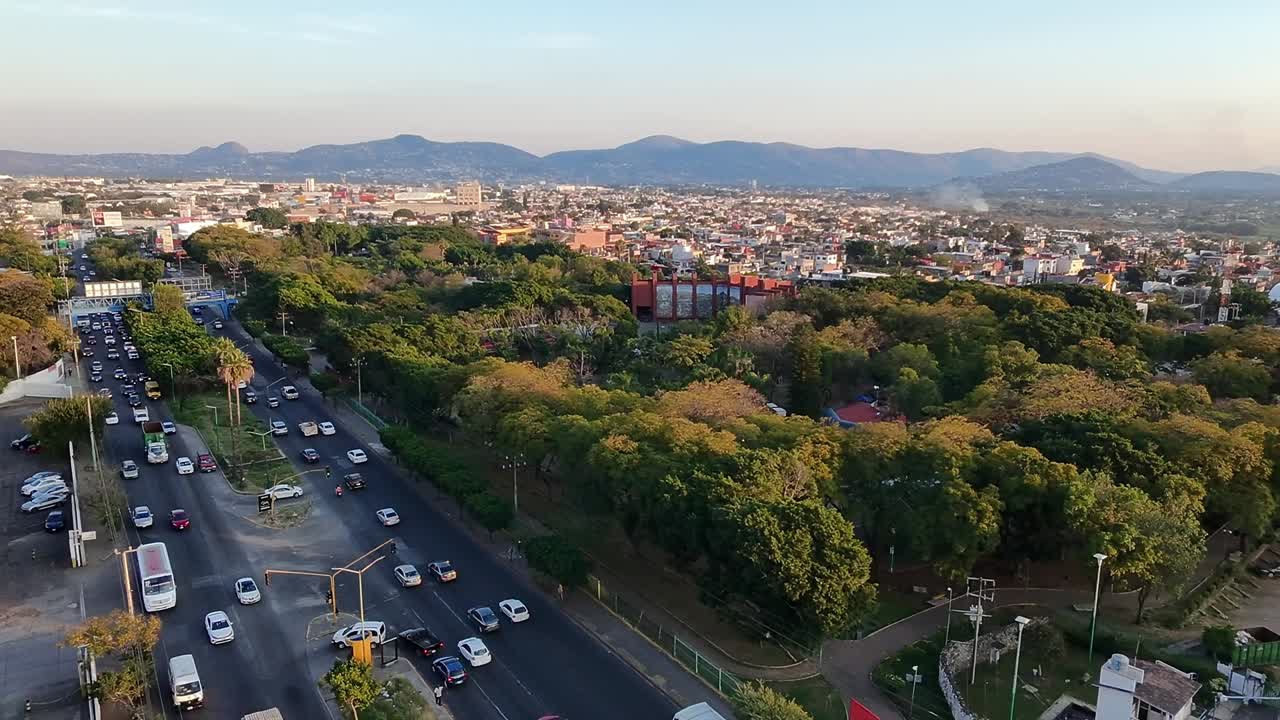 Aerial view capturing traffic flowing near Alameda Park in Cuernavaca, Morelos, Mexico, during a vibrant sunset, with majestic mountains rising in the background