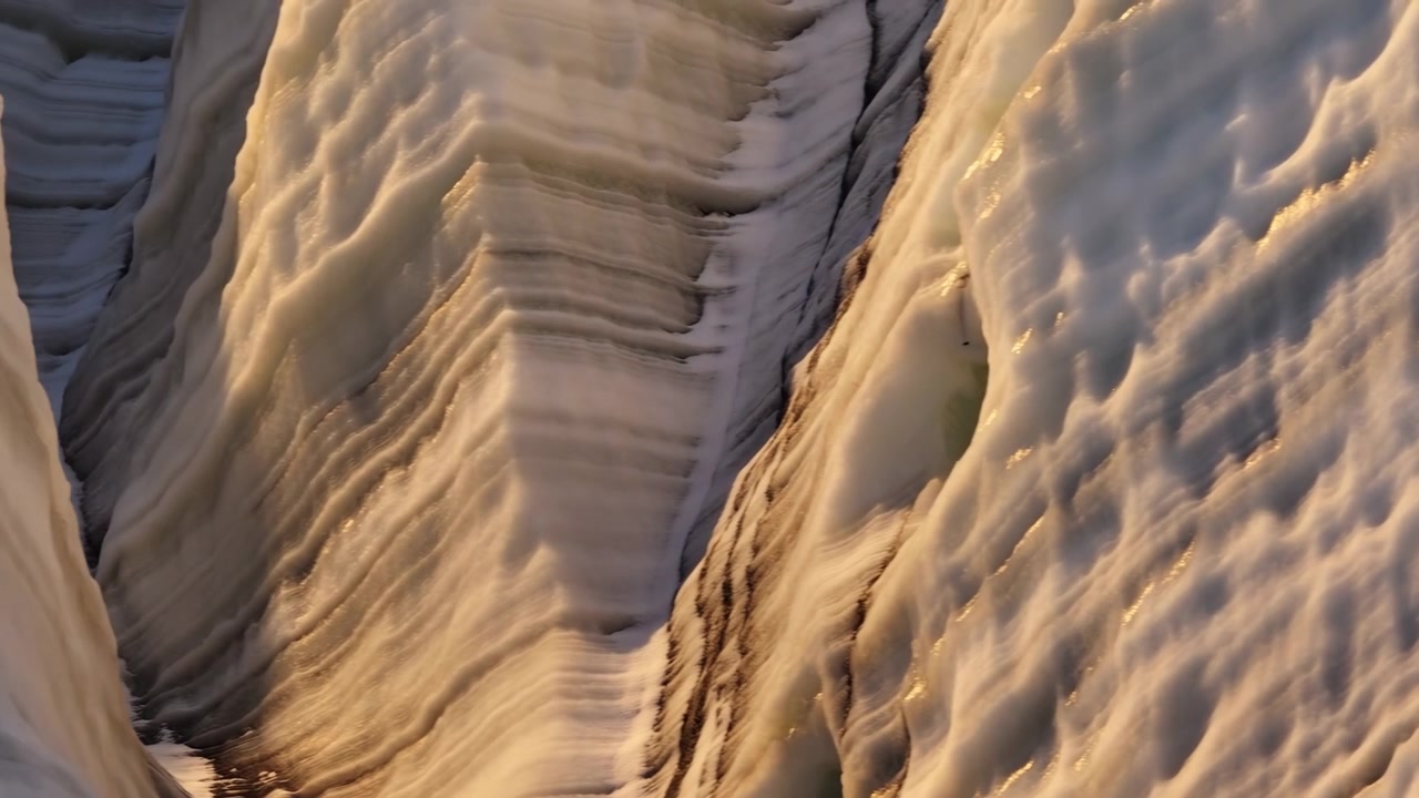 Close oblique view into narrow glacier crevasse flanked by steep layered ice walls, scalloped snow faces, translucent frost textures, deep shadow, and warm low-angle sunlight across rugged alpine