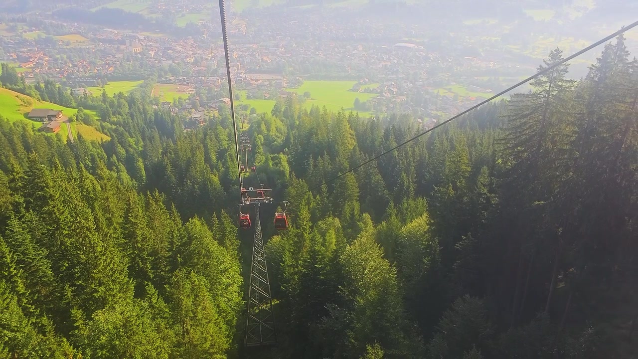 Ampia vista di Kitzbühel, Austria, dalla cabinovia, che mostra la città sottostante, una pineta, prati ondulati, case alpine e le Alpi tirolesi stratificate in una località turistica sotto il caldo sole estivo.