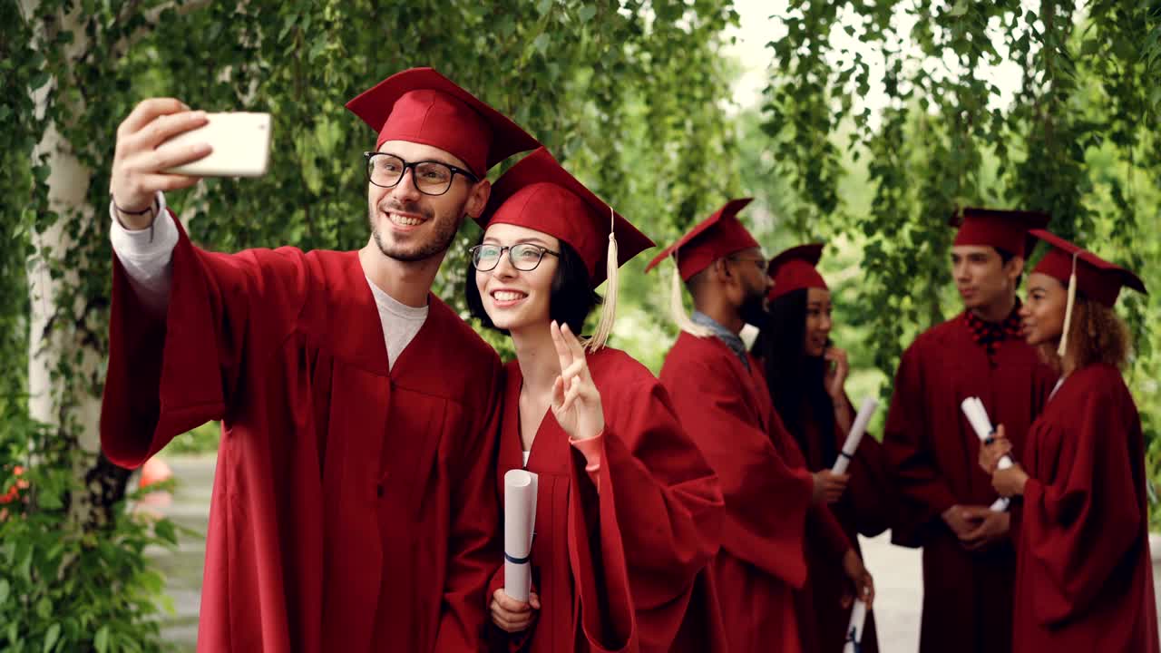 Cheerful couple of graduating students is taking selfie using smartphone, young man and woman are holding diplomas, looking at smart phone camera and smiling.