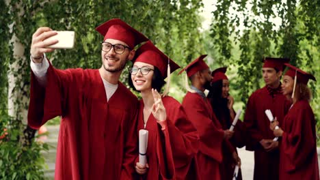 cheerful couple of graduating students is taking selfie using smartphone, young man and woman are holding diplomas, looking at smart phone camera and smiling.