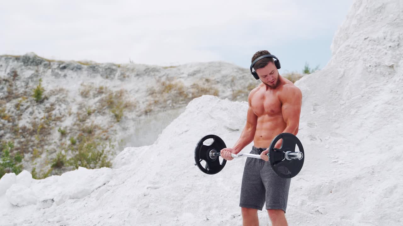 Athletic man with a naked torso in headphones does exercises for the biceps barbell. Fitness in nature